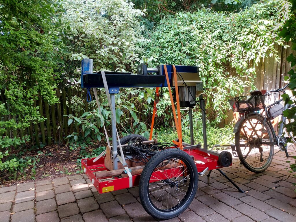 Fahrrad mit Anhänger. Auf dem Anhänger ein Holzspalter verzurrt mit Spanngurten. Unter dem Spalter eine Werkzeugkiste, Verlängerungskabel, Hammer und Axt