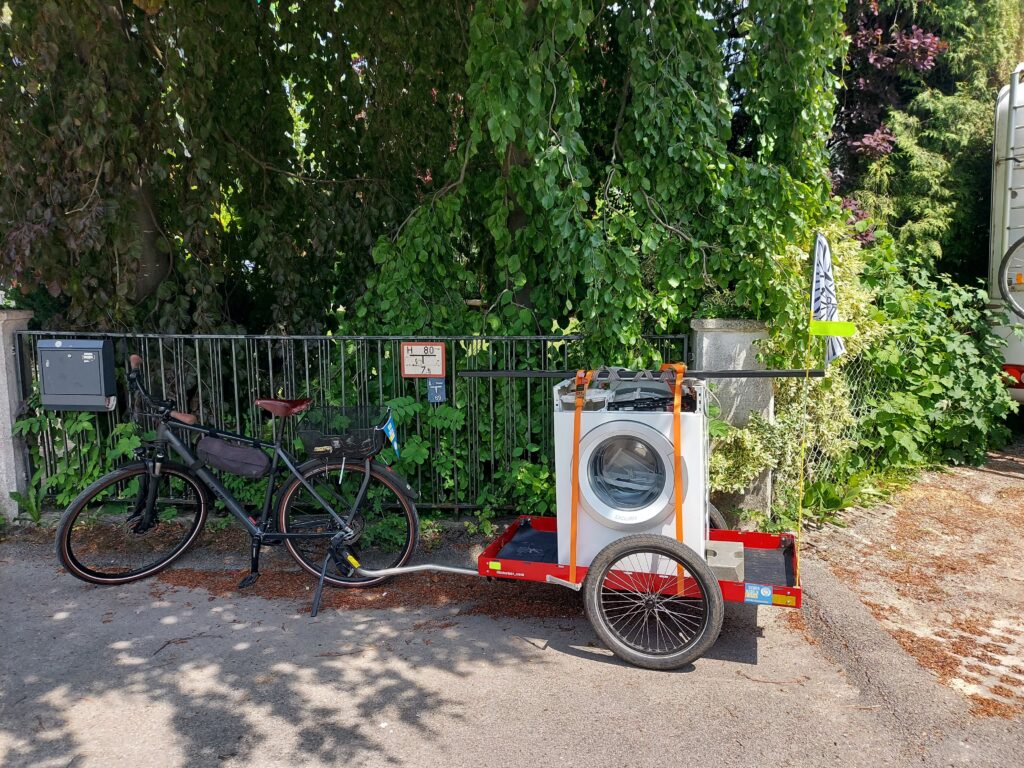 Fahrrad mit Anhänger vor Gartenzaun. Auf dem Anhänger eine Waschmaschine, befestigt mit Spanngurten.